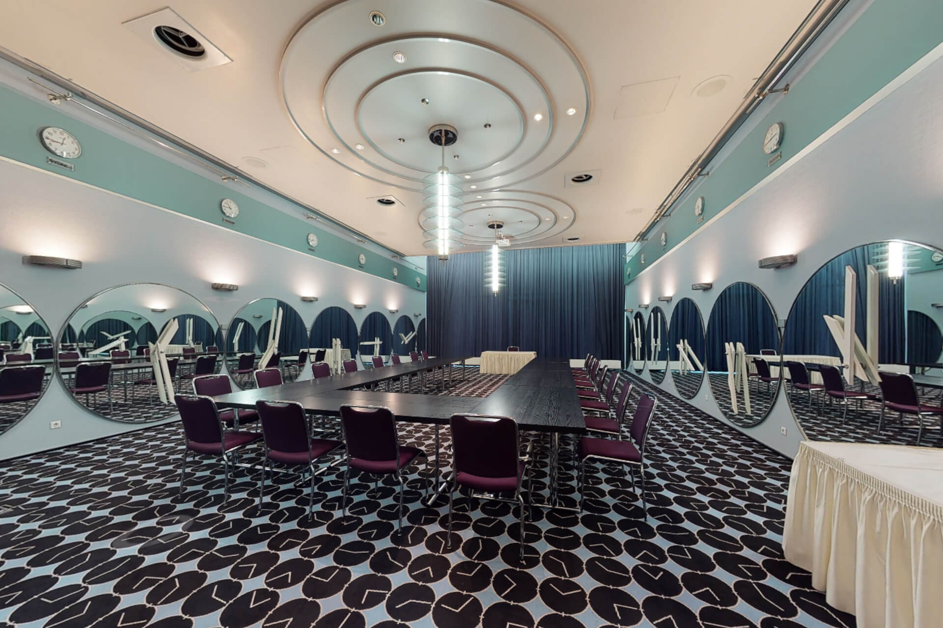 Ein Blick in den Salon Danzig, eines der Konferenz-Zimmer bei CONGRESS BREMEN. Tische sind als aufgebaut, Stühle herum drapirt. Der Fußboden hat ein schwarz-weißes Muster mit Kreiselementen, die sich auch am Stuck der Decke wiederfinden. Farblich bestimmt gedecktes Blau bis Türkis mit Schwarz am Boden und Beige an der Decke.
